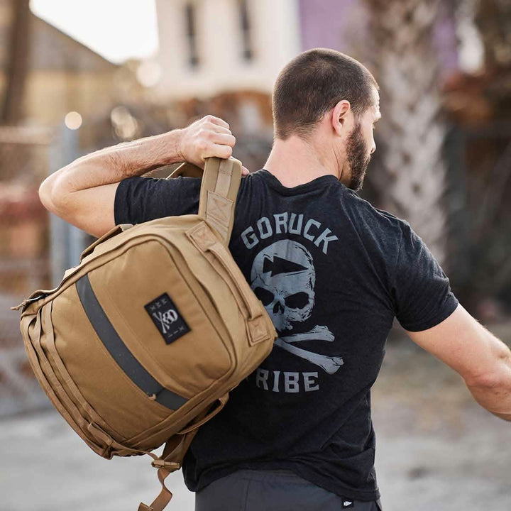 A bearded man carrying a tan backpack over his shoulder is wearing the GORUCK Tribe Tee - Tri-Blend from vendor-unknown, featuring a skull and crossbones design on the front. The bold "GORUCK Tribe" text across the back symbolizes shared pain and camaraderie associated with GORUCK Challenges, all set against an outdoor urban backdrop.