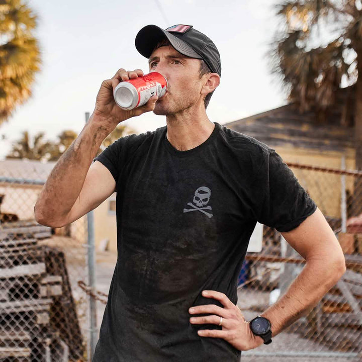A man wearing a GORUCK Tribe Tee - Tri-Blend and a black cap enjoys a refreshing drink from a soda can outdoors, embodying the spirit of the GORUCK Challenge. Standing next to a chain-link fence with one hand on his hip, he's surrounded by trees and an aged building—a scene capturing shared pain and camaraderie in nature's embrace.