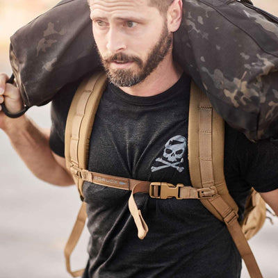 A man participates in the GORUCK Challenge, shouldering a hefty black sandbag. He's wearing the GORUCK Tribe Tee - Tri-Blend, which features a white skull and crossbones design. Along with this, he carries a tan backpack with buckles. His expression is focused and determined, as he embraces the shared challenge alongside fellow participants.