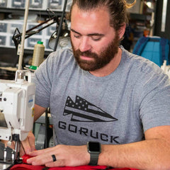 Sporting a gray GORUCK USA Tee - Tri-blend, the bearded man expertly sews red fabric on a sewing machine in his workshop, demonstrating his Special Forces precision and skill.