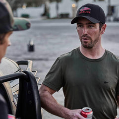 Man in GORUCK cap and green shirt holding a drink, outdoors by a vehicle