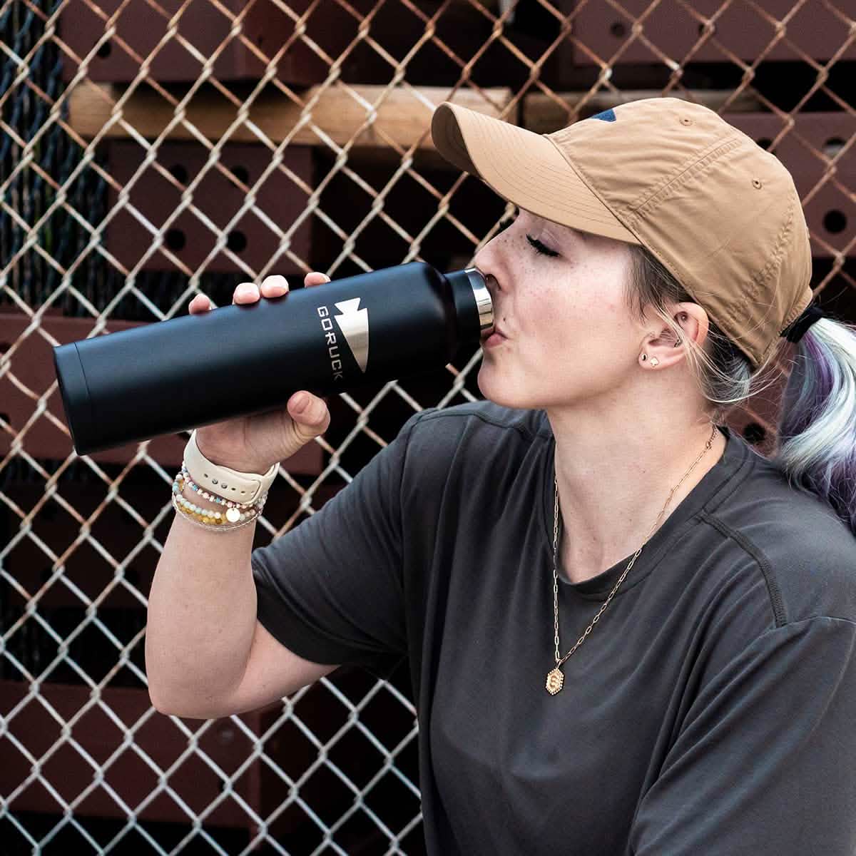 A person wearing a beige cap and gray shirt drinks from a GORUCK x RTIC double wall insulated black water bottle while sitting in front of a chain-link fence. They have light hair tied back and wear several bracelets and a necklace.