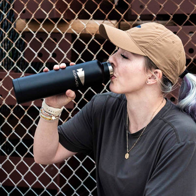 A person wearing a beige cap and gray shirt drinks from a GORUCK x RTIC double wall insulated black water bottle while sitting in front of a chain-link fence. They have light hair tied back and wear several bracelets and a necklace.