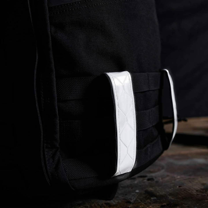 A close-up of a black GORUCK backpack showcases the Reflective Ruck Bands, which feature a diamond pattern in white to enhance nighttime visibility and add stylish contrast to the bag's smooth fabric. The dark wooden background beautifully highlights the bag’s details.