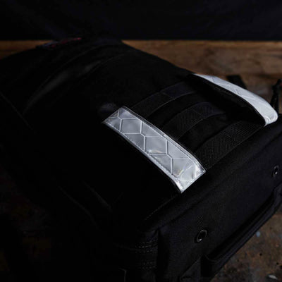 A black backpack with a luxurious quilted texture and gleaming silver straps is enhanced by GORUCK's Reflective Ruck Bands for improved nighttime visibility. The bag, which rests on a wooden surface against a dark background, boasts grommet details and exudes both durability and style.