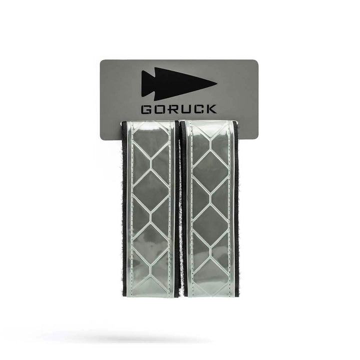 The GORUCK Reflective Ruck Bands, showcasing a diamond pattern, are affixed to a gray card emblazoned with the arrowhead logo and the brand name. Equipped with MOLLE webbing for secure attachment, these bands improve nighttime visibility, enhancing safety during outdoor activities.