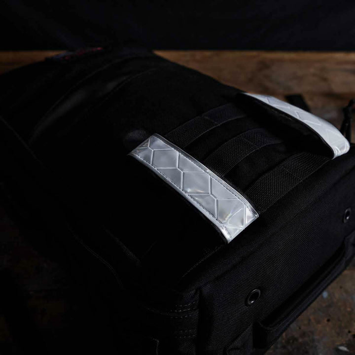 A black backpack with a luxurious quilted texture and gleaming silver straps is enhanced by GORUCK's Reflective Ruck Bands for improved nighttime visibility. The bag, which rests on a wooden surface against a dark background, boasts grommet details and exudes both durability and style.